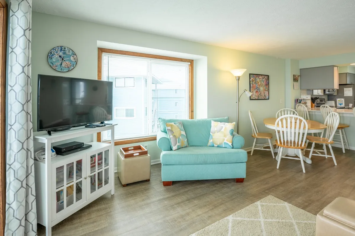 A bright, open-concept living and dining area with a turquoise sofa, a dining table, a large TV unit, and an expansive window that bathes the space in natural light. The room is set up for both lounging and gathering, echoing the theme of shared experiences.