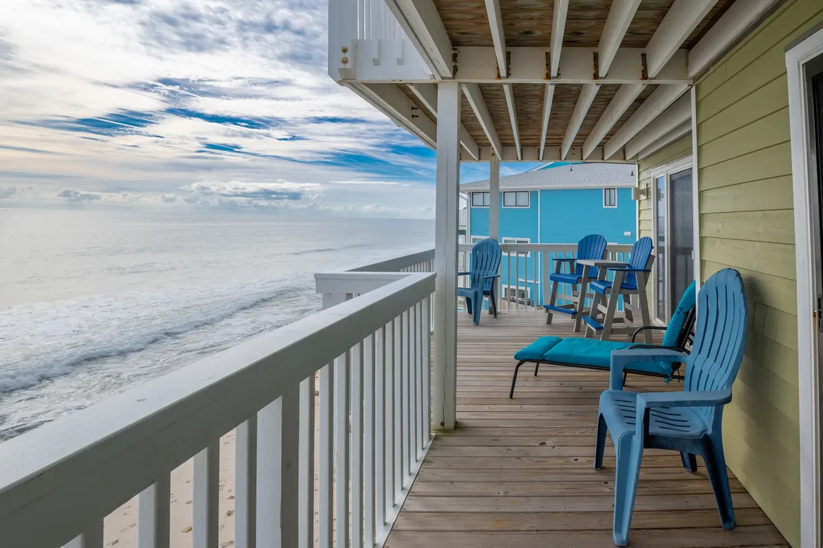 A tranquil oceanfront deck with blue Adirondack chairs and a wooden railing, offering a panoramic view of the sea. The deck feels inviting and perfect for relaxing, reading, or socializing while enjoying the sound and sight of the ocean.