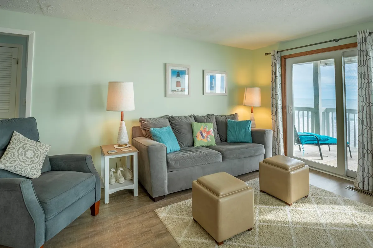 This inviting living space features a plush sofa adorned with decorative pillows, side lamps, attractive artwork, and a sliding glass door that leads to an oceanfront view. The ambiance is cozy and cheerful, perfect for relaxing, socializing, or enjoying the morning sunrise from indoors.