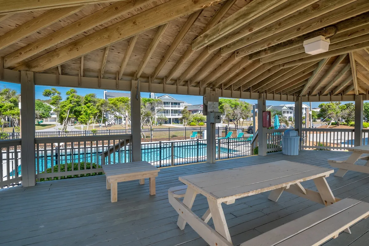 A covered outdoor picnic area with wooden beams, featuring picnic tables and a view of the swimming pool beyond. The setting is framed by greenery, suggesting a relaxed, social environment for families and groups to gather outdoors after a day of adventure.
