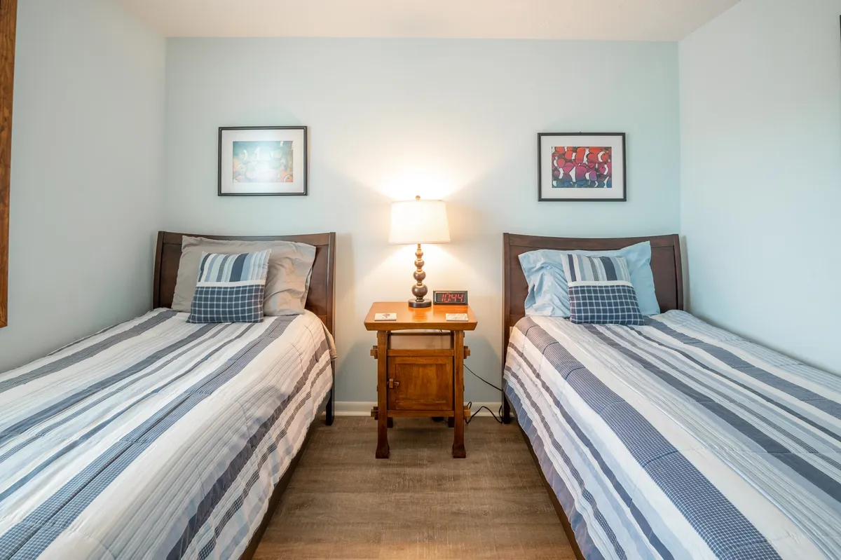 A cozy, friendly second bedroom with two twin beds separated by a nightstand and lamp, complemented by framed artwork on the wall. Warm lighting and thoughtful decor create an inviting, comfortable space ideal for children or friends.