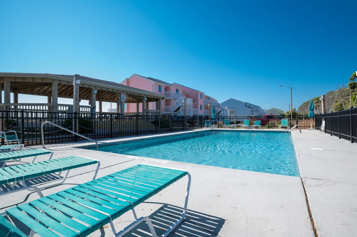 A lively resort swimming pool scene surrounded by lounge chairs and adjacent buildings, suggesting a vibrant, family-friendly atmosphere. The setup hints at opportunities for swimming, sunbathing, and children playing while parents relax.