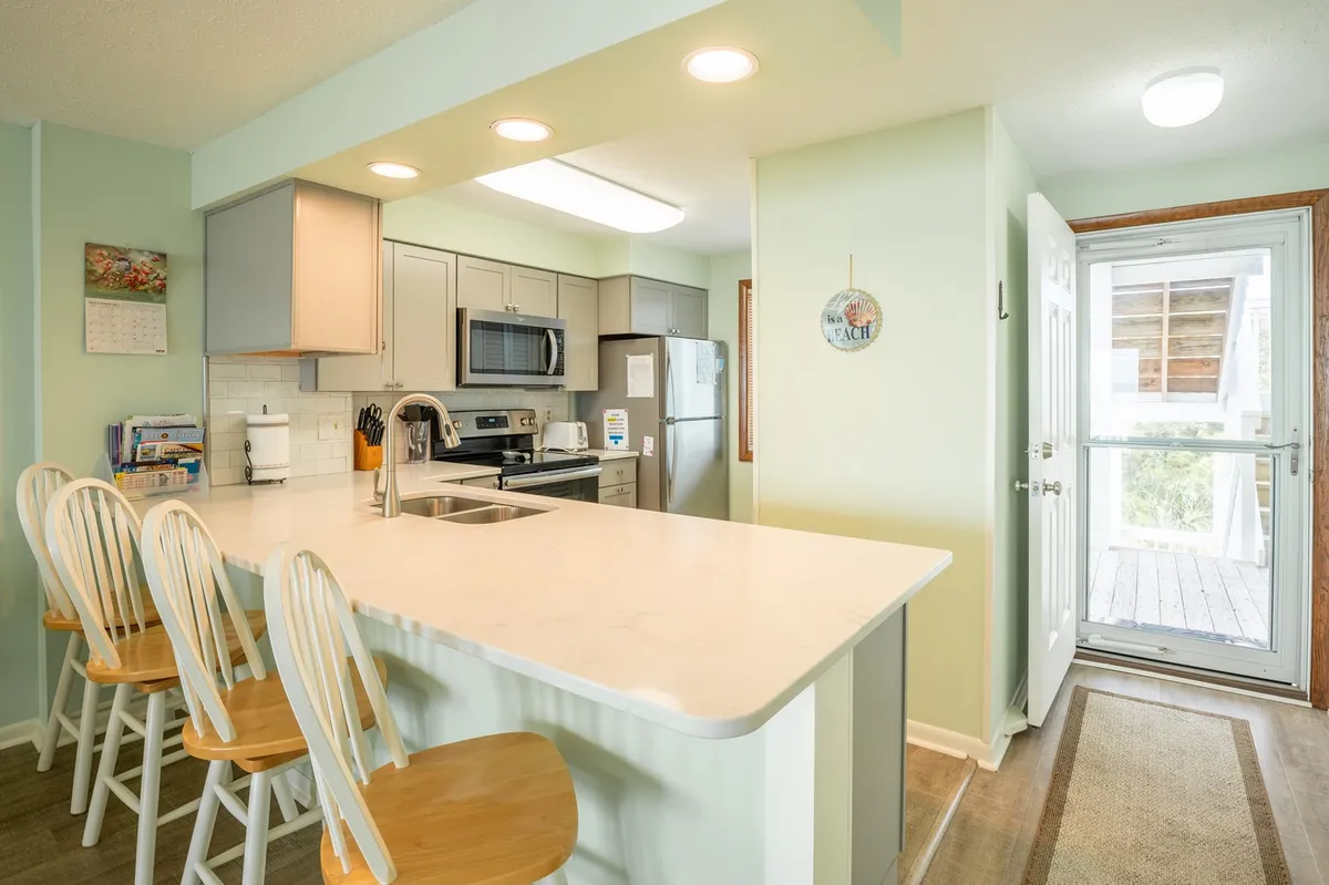 This image captures a welcoming kitchen with a central island, bar stools, visible kitchen appliances, and abundant natural light streaming in from a nearby door. The layout highlights the kitchen bar seating area, modern cabinetry, and a bright, functional gathering space.