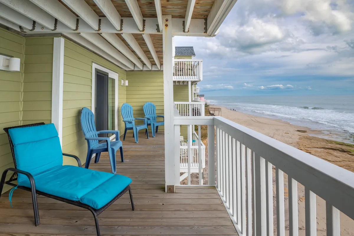 A tranquil balcony with a blue lounge chair offers a sweeping beach view framed by a wooden railing. The scene invites moments of relaxation, sunrise watching, or sharing stories with friends after a day of exploring.