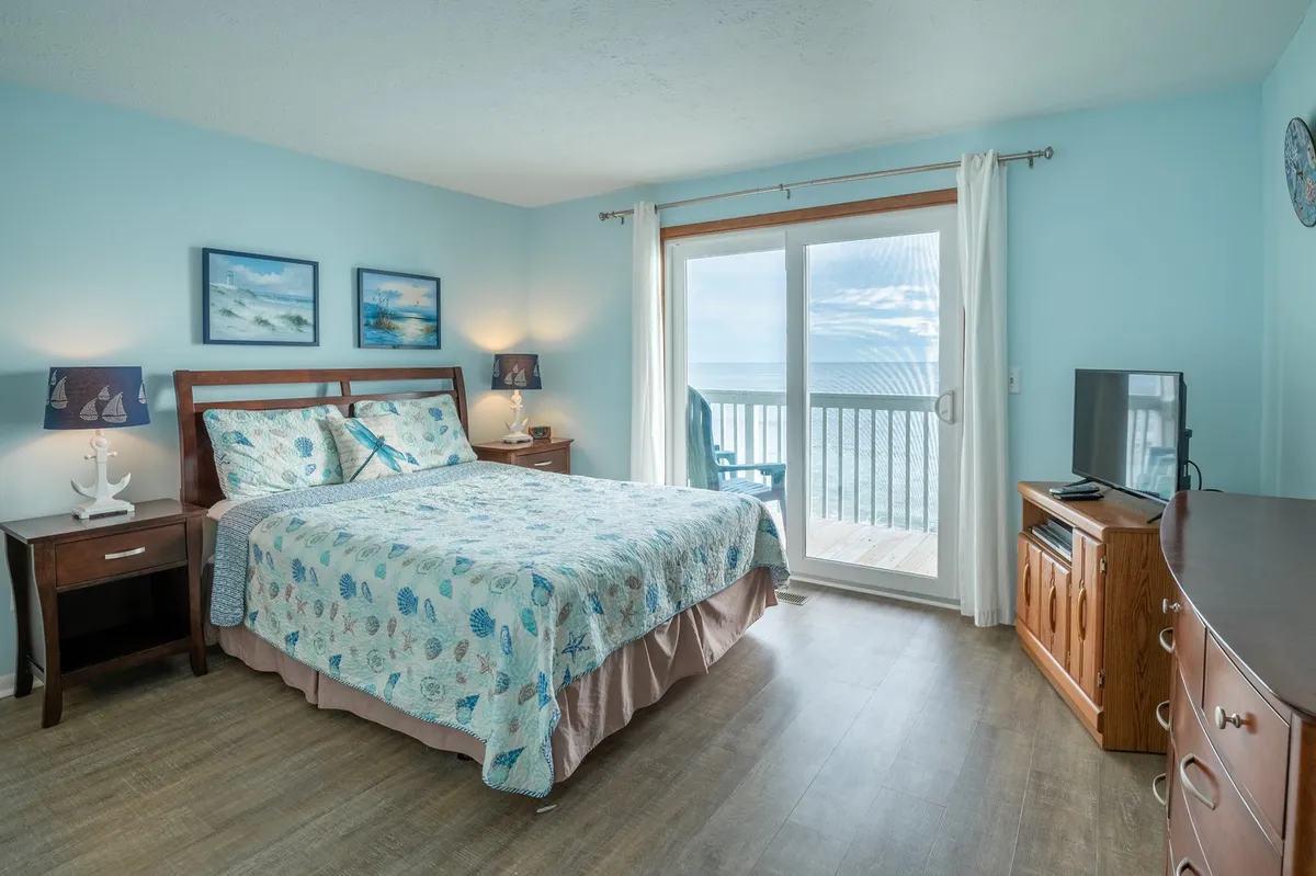 A serene primary bedroom with a queen-sized bed covered in a patterned quilt, large sliding glass doors opening to a balcony, nautical-themed artwork on the walls, and wooden bedside tables with lamps. A TV is also visible, enhancing the room's comfort. The space is bright and airy, with direct ocean views creating a tranquil, inviting retreat.