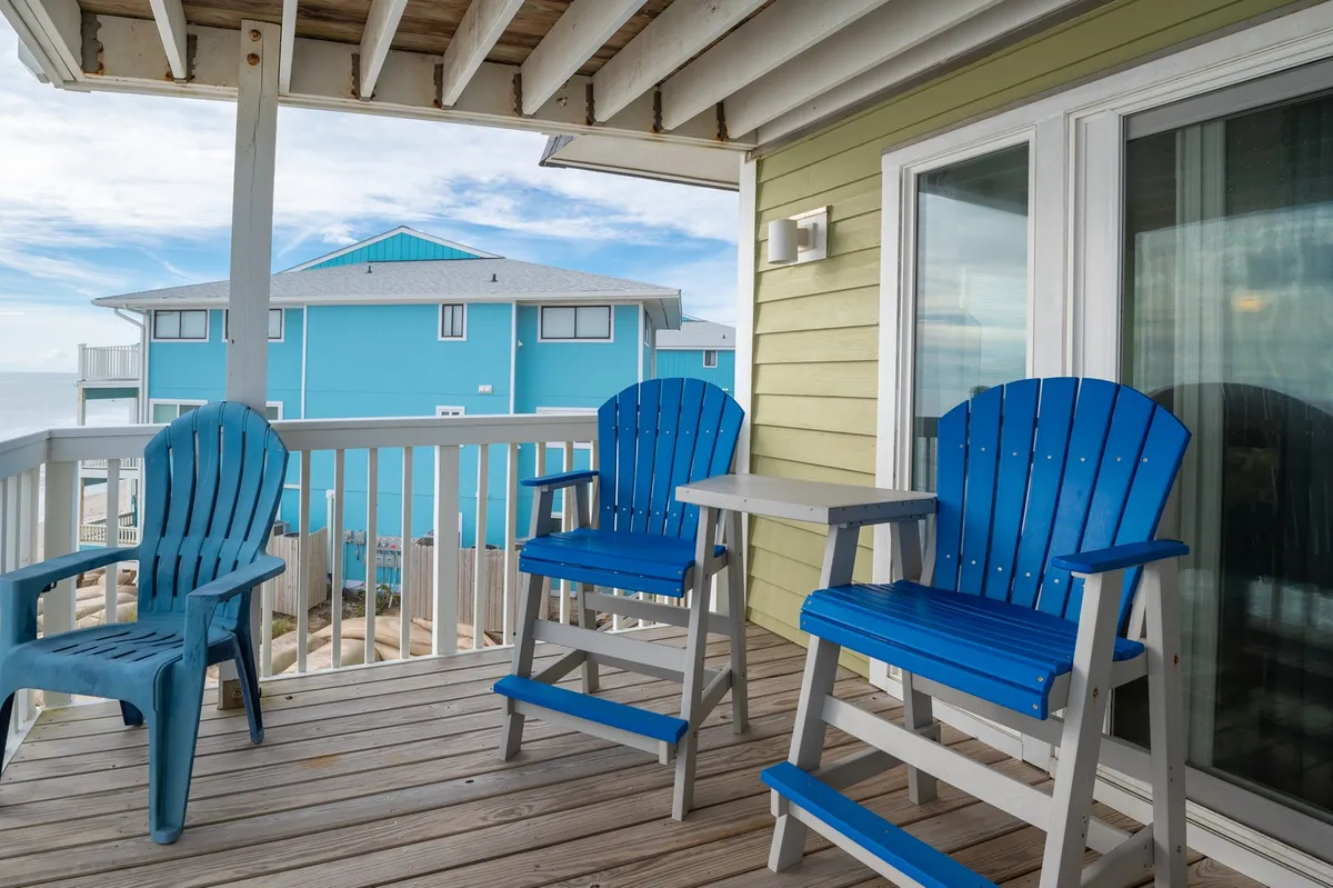 A cozy outdoor setting featuring blue Adirondack chairs and a wooden table on a private balcony, overlooking a neighboring blue building. The tranquil atmosphere invites guests to relax, sip coffee, and take in sweeping views, with the potential for sunrise ocean vistas.