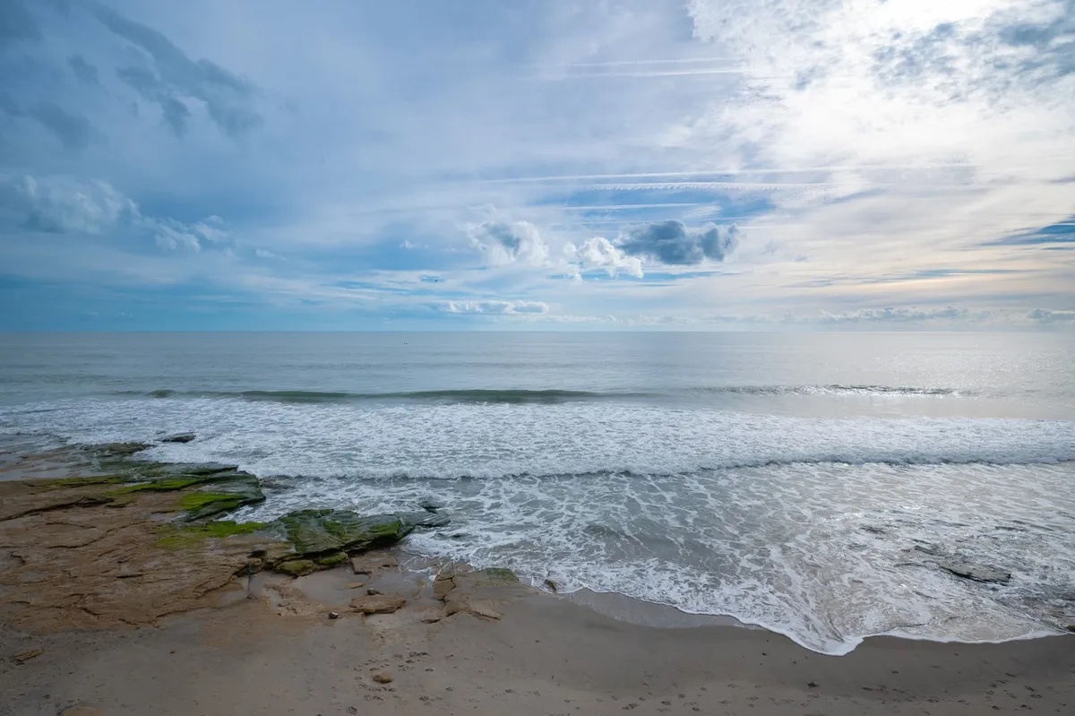 A sweeping view of the natural shoreline, featuring gentle ocean waves rolling onto a rocky shore. The foreground shows the distinctive coquina rock formations, while the distant horizon and moody cloud formations evoke a sense of tranquility and awe. There are no buildings or people, emphasizing the untouched beauty and wild nature of the setting.